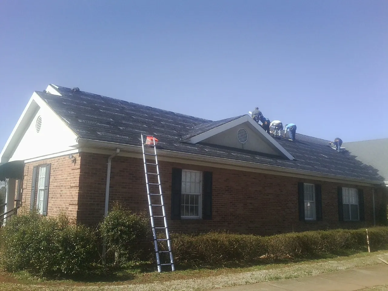 Crew tearing shingles off a brick ranch roof with a ladder set — Emergency Roof Repair work by our crew in Gaston